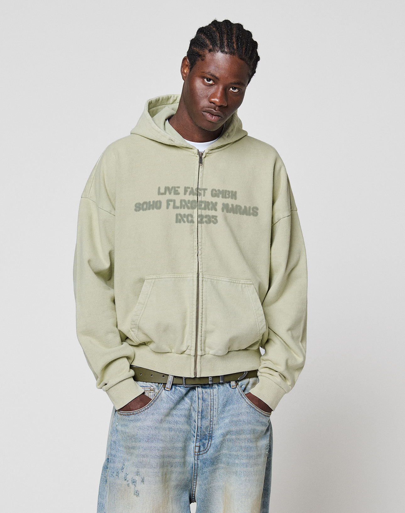 A person with braided hair wears the LIVE FAST DIE YOUNG LFDY District Hooded Zip in light green with a faded frontprint and light blue baggy jeans, standing hands-in-pockets against a white background. The hoodie is made from 100% Baumwolle.