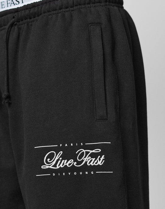 Close-up of Wide Cuff Jogger by LIVE FAST DIE YOUNG in black French Terry. Features white embroidered "LIVE FAST DIE YOUNG" text near the pocket, with visible elastic waistband and pocket opening.