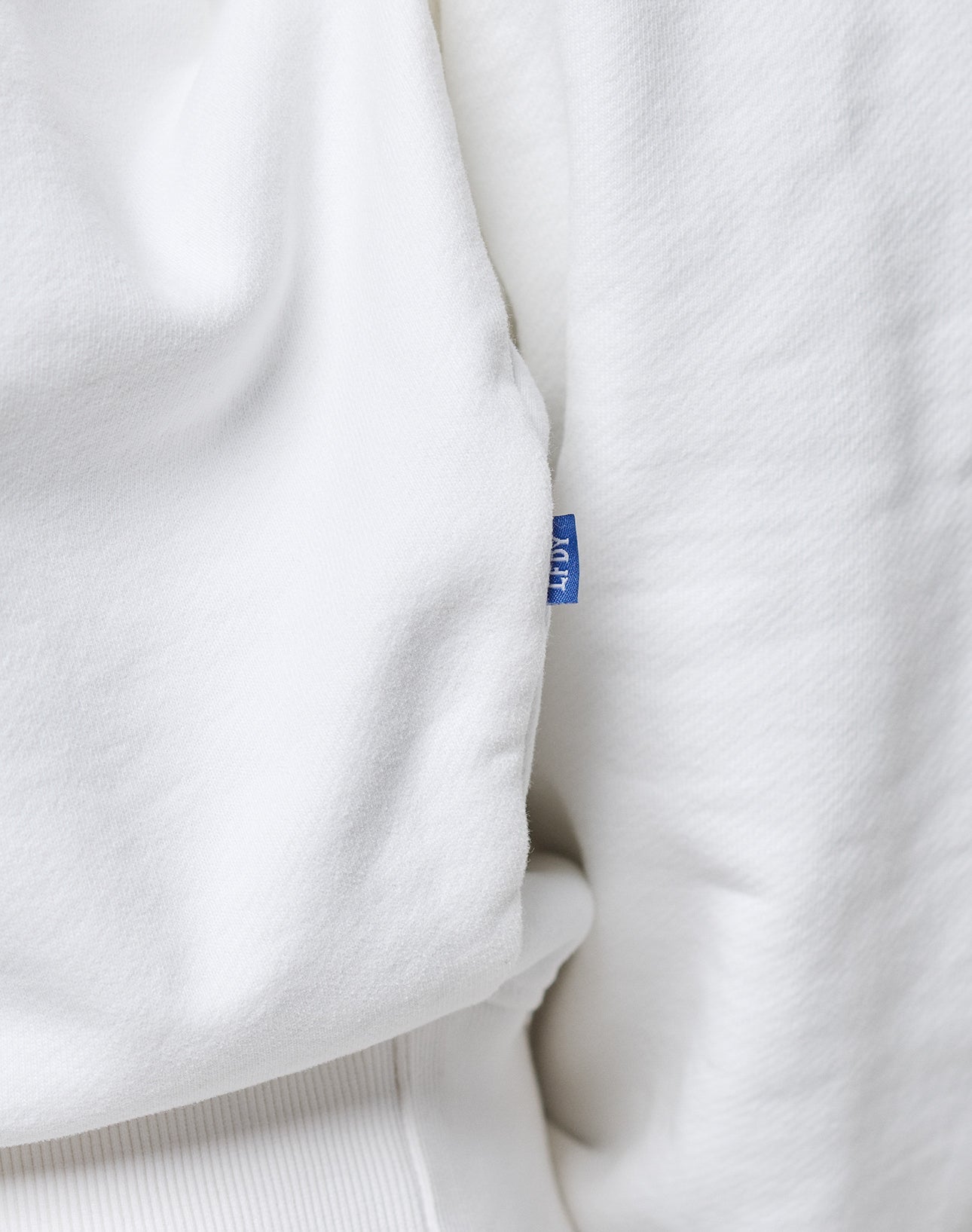 Close-up of an LFDY Radiator Sweater by LIVE FAST DIE YOUNG in white Baumwolle Polyester, featuring a small blue tag with partial white text sewn into the seam. The garment’s texture and subtle folds are clearly visible.