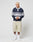 A person with short blond hair wears the LIVE FAST DIE YOUNG LFDY Striped Polo in navy and gray, beige knee-length cotton twill shorts, white socks, and white sneakers, standing against a plain light background.