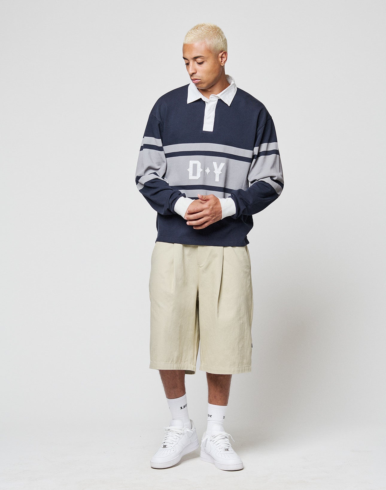 A person with short blond hair wears the LIVE FAST DIE YOUNG LFDY Striped Polo in navy and gray, beige knee-length cotton twill shorts, white socks, and white sneakers, standing against a plain light background.
