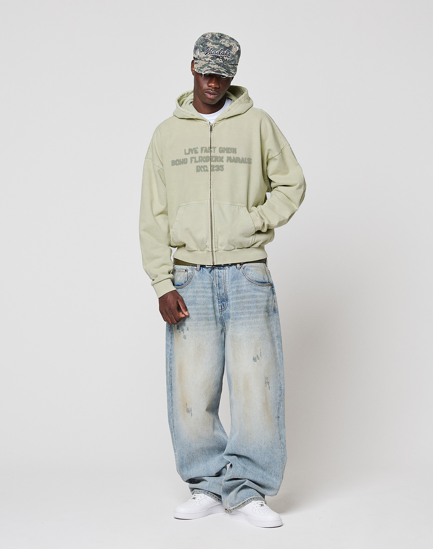 A person wears a patterned cap, LFDY District Hooded Zip by LIVE FAST DIE YOUNG in light green with front print, loose faded blue jeans, and white sneakers, standing against a plain white background.