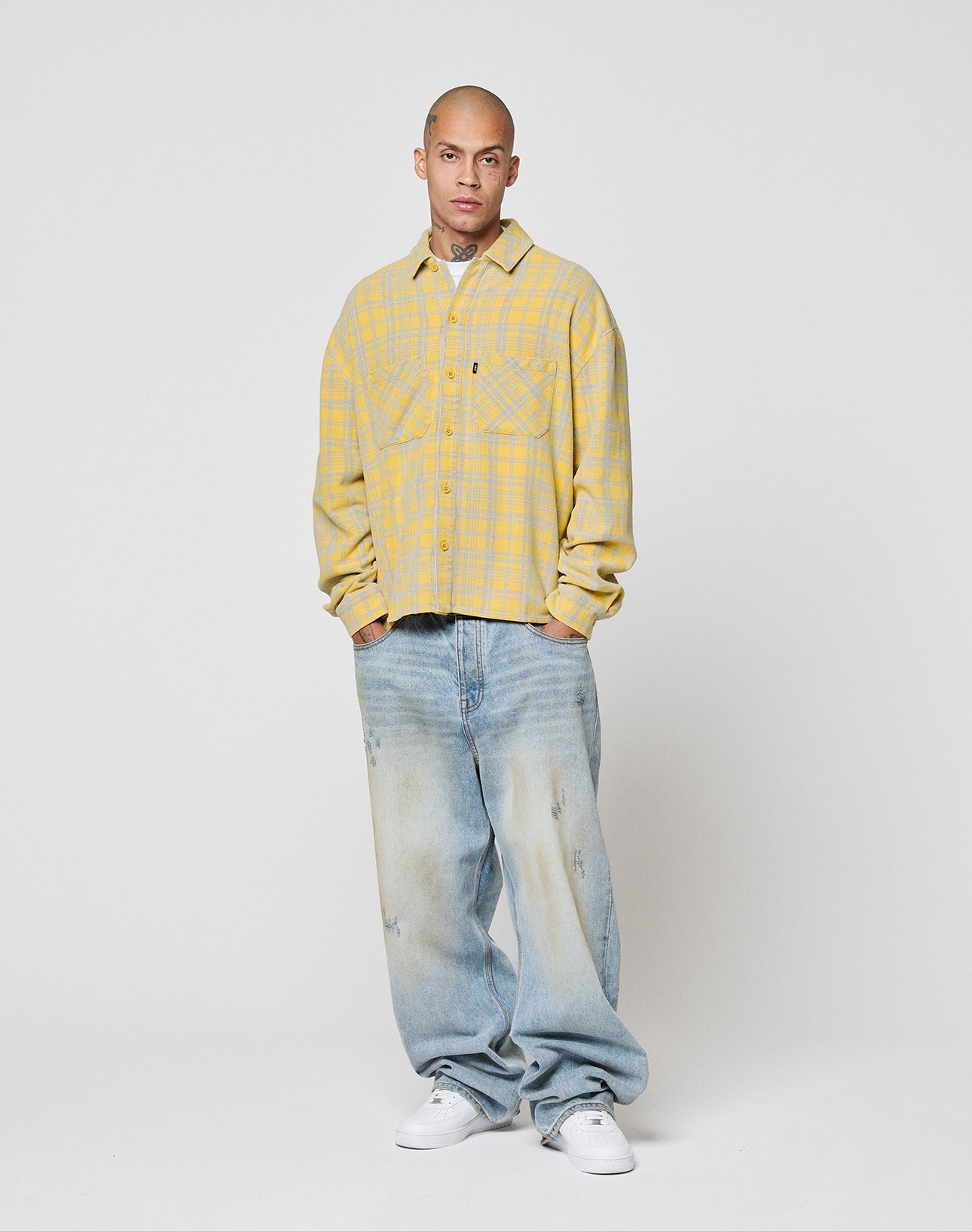 Against a plain white background, a person with neck tattoos and a shaved head wears the LIVE FAST DIE YOUNG Vintage Flannel in yellow plaid, 100% Baumwolle, light blue baggy jeans with Vintage Waschung, and white sneakers. Hands in pockets.