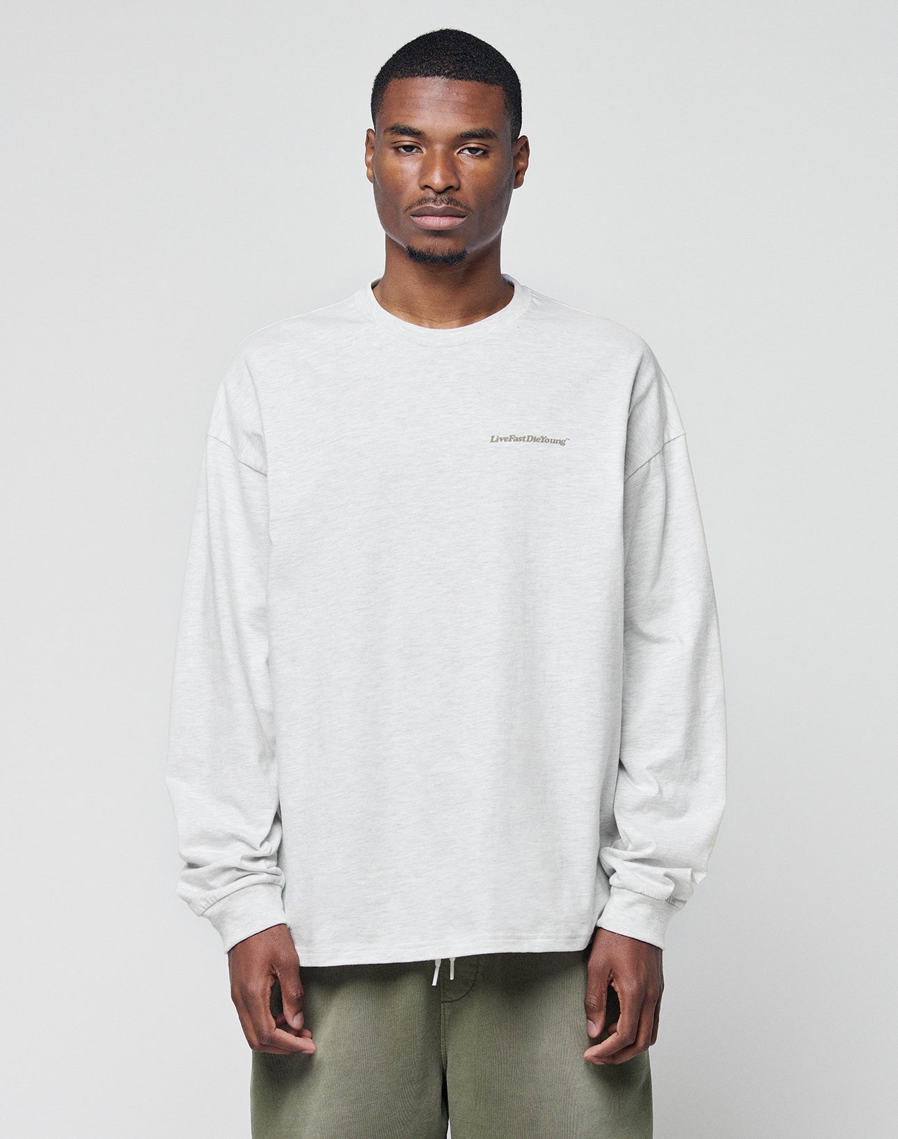 A man with short hair wears the LIVE FAST DIE YOUNG Basic Longsleeve, a light gray, logo-print, oversize sweatshirt, paired with olive green pants, standing against a plain light background. | model