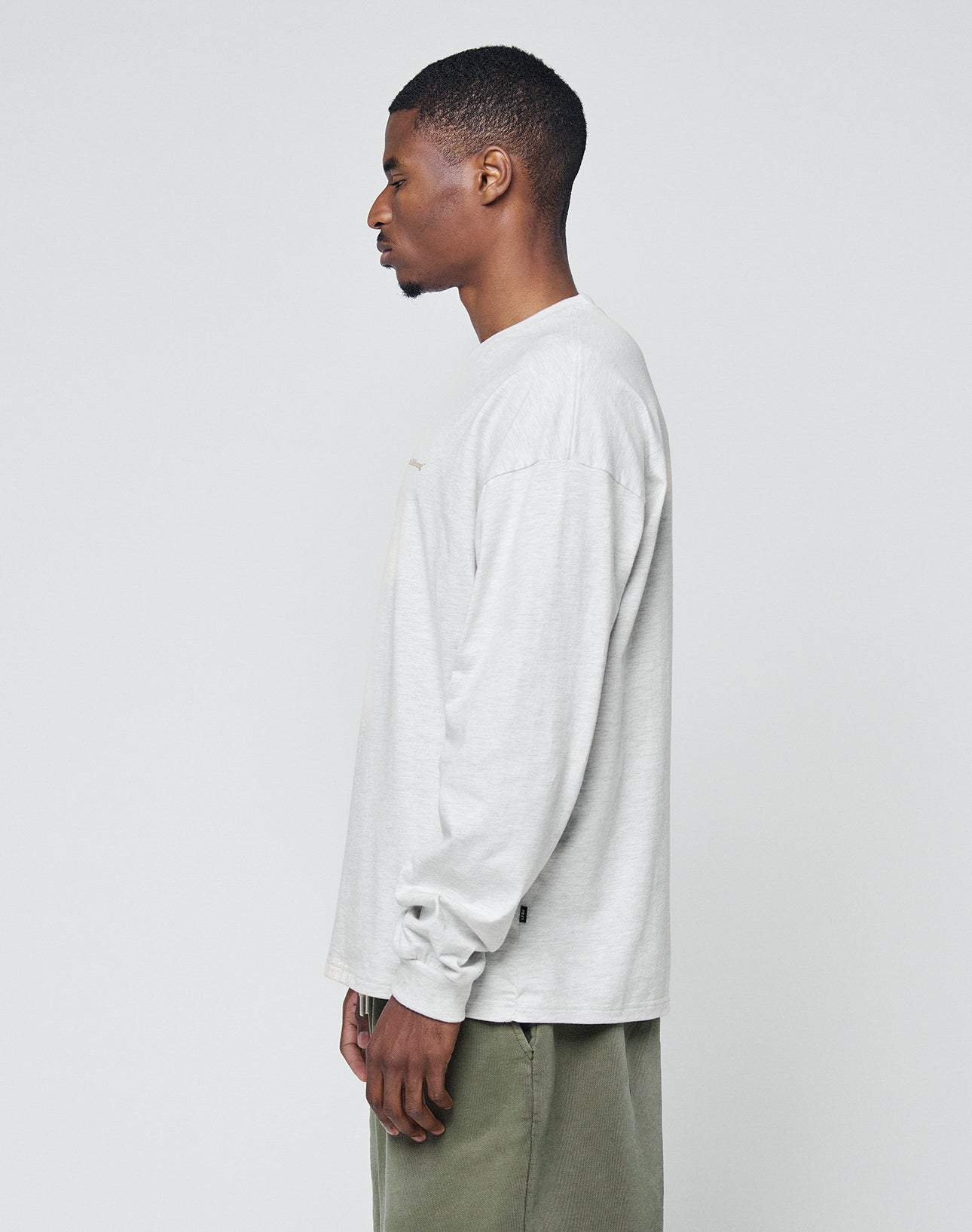 A man stands in profile against a plain light background, wearing the LIVE FAST DIE YOUNG Basic Longsleeve in light gray 100% cotton and olive green pants.