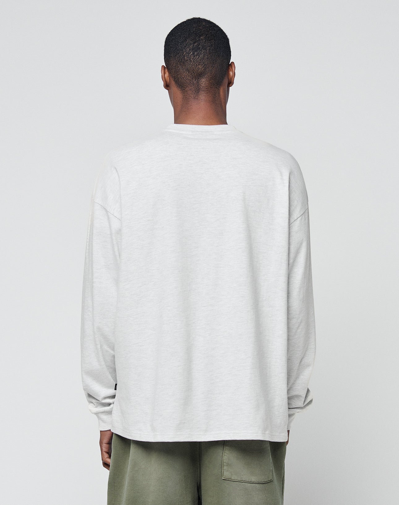 A person with short hair, facing away from the camera, wears a LIVE FAST DIE YOUNG Basic Longsleeve in light gray (100% Baumwolle) and olive green pants against a plain light background.