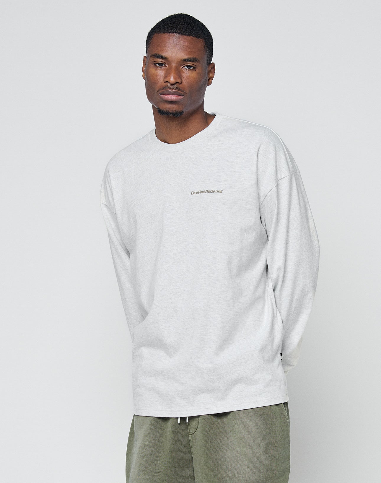 A man in a light gray LIVE FAST DIE YOUNG Basic Longsleeve made from 100% Baumwolle and olive green pants stands against a plain background, calmly facing the camera.