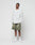 Against a plain white backdrop, a man wears the LIVE FAST DIE YOUNG Basic Longsleeve in light gray with an oversize fit, styled with olive green shorts, white crew socks, and white sneakers. His hands are in his pockets and he has a neutral expression.