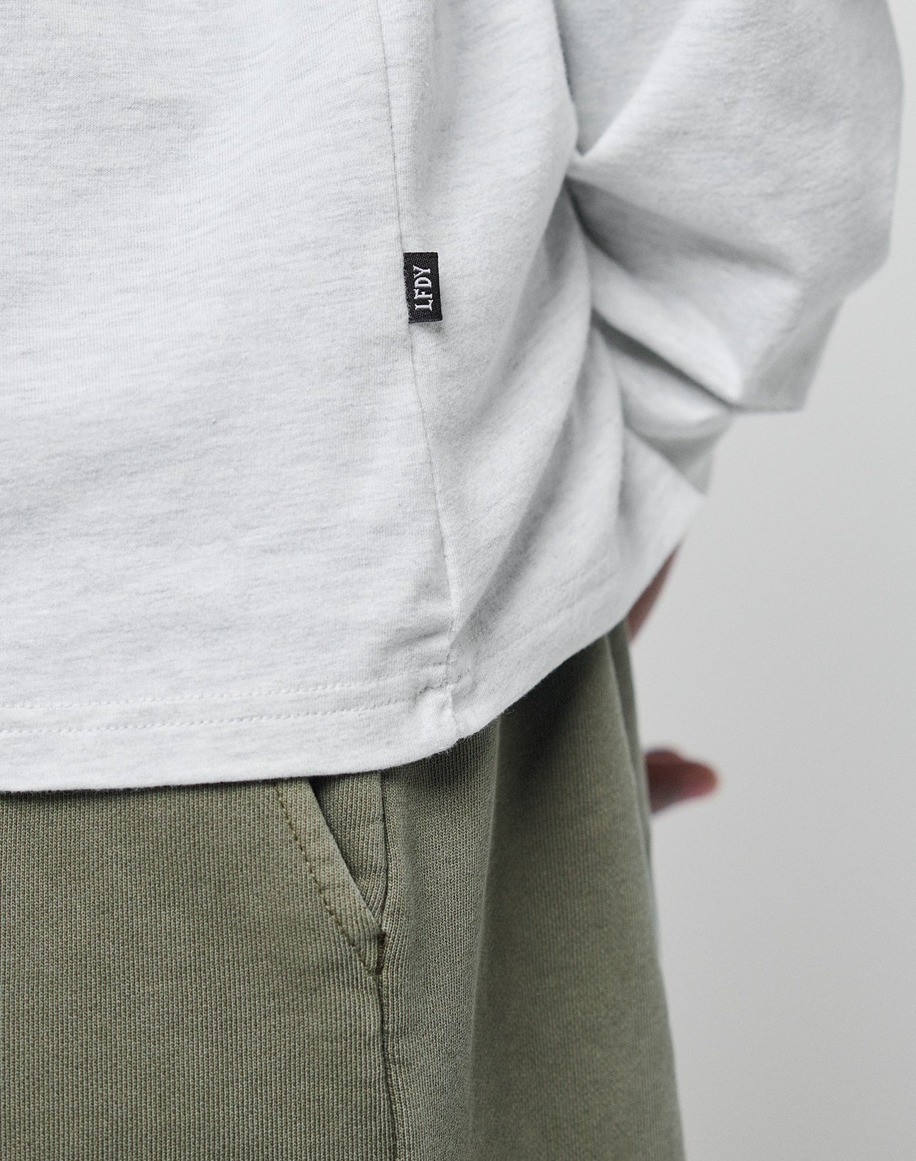 Close-up of a person in an oversize fit white LIVE FAST DIE YOUNG Basic Longsleeve (100% Baumwolle) with a small black IFPY tag and subtle logo-print, paired with olive green pants; the persons arm is bent behind their back.