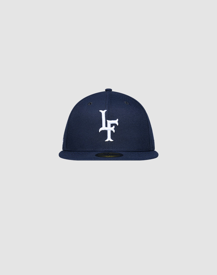 The LFDY x New Era 59Fifty Paris Fitted Cap by LIVE FAST DIE YOUNG features a navy blue design with white LF embroidery on the front and a flat brim, displayed on a plain light gray background. Größentabelle available for sizing.| plain