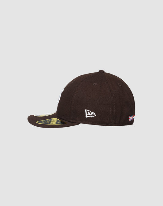 The LFDY X NEW ERA 59FIFTY LOW PROFILE by LIVE FAST DIE YOUNG is a black baseball cap with a curved brim and stickers, a white side logo, and a small flag above the adjustable back strap.