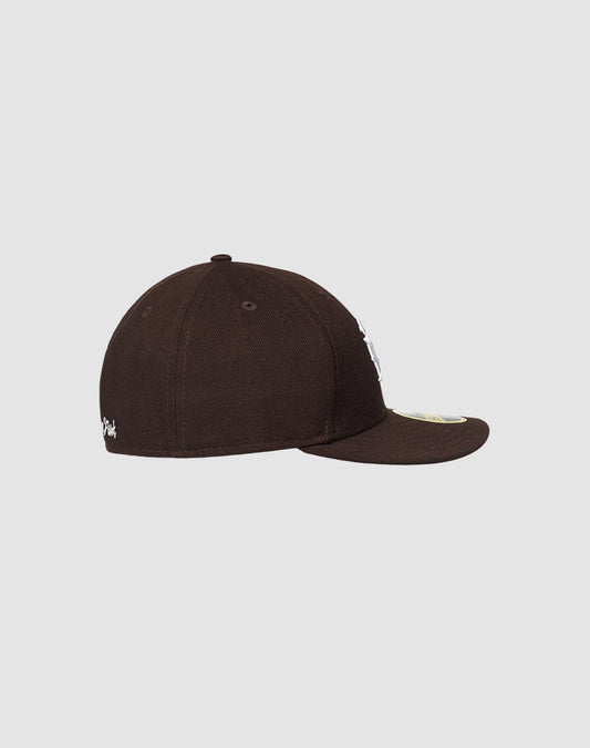 The LFDY x NEW ERA 59FIFTY Low Profile by LIVE FAST DIE YOUNG is shown from the side with a flat brim and sticker, its dark brown color standing out against a plain light gray background.