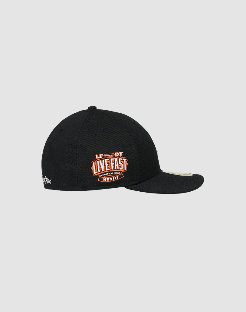 The LFDY X NEW ERA 59FIFTY LOW PROFILE by LIVE FAST DIE YOUNG is a black fitted cap with an orange and white LIVE FAST patch on the side and small white text on the back, shown on a light gray background.