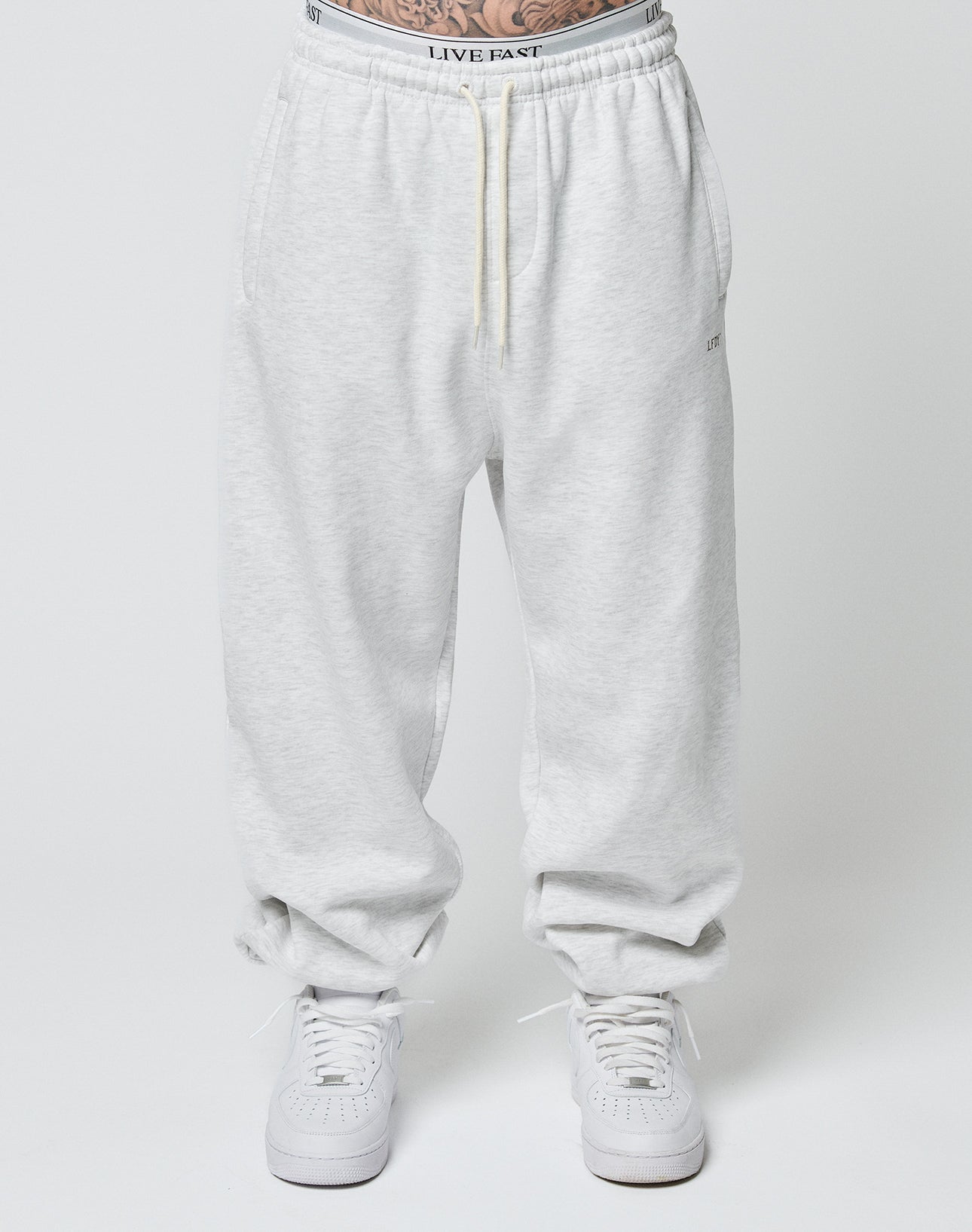 A person wears Basic Loose Jogger sweatpants by LIVE FAST DIE YOUNG in light gray with a loose fit and elastic waistband, paired with white sneakers. The visible waistband reads LIVE FAST. Background is plain white.