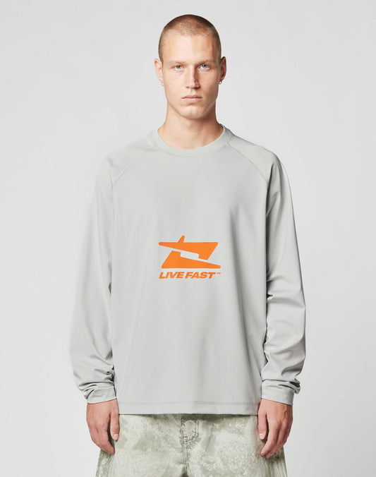 A person with short hair wears the LFDY Sports Longsleeve by LIVE FAST DIE YOUNG, featuring Raglan sleeves and an orange LIVE FAST graphic on light gray, paired with light-colored pants against a plain background.| model