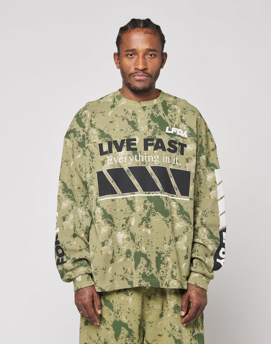 A man with braided hair, beard, and mustache wears the LIVE FAST DIE YOUNG LFDY Racing Waffle Longsleeve—a green oversized camo sweatshirt with bold LIVE FAST Everything in it. front print and black/white graphics—and matching pants.