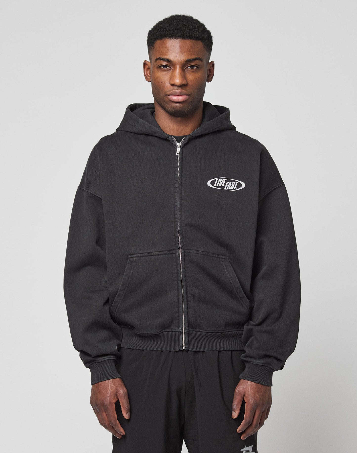 Facing forward against a light gray background, a person wears the LFDY Canine Hooded Zip by LIVE FAST DIE YOUNG—an oversize fit black zip-up hoodie with a small silver LIVE FAST logo on the left chest—paired with black pants.
