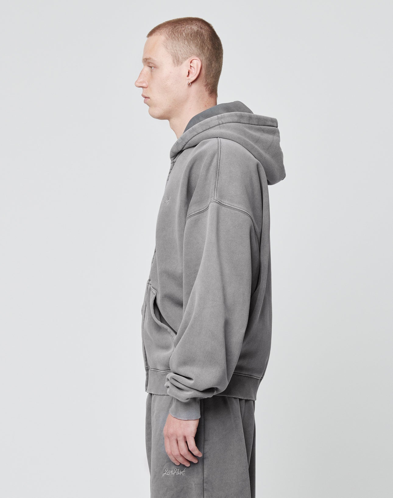 A man with short hair stands in profile, dressed in the LIVE FAST DIE YOUNG Basic Script Hooded Zip and matching gray cotton sweatpants with a subtle vintage wash, set against a plain light background.