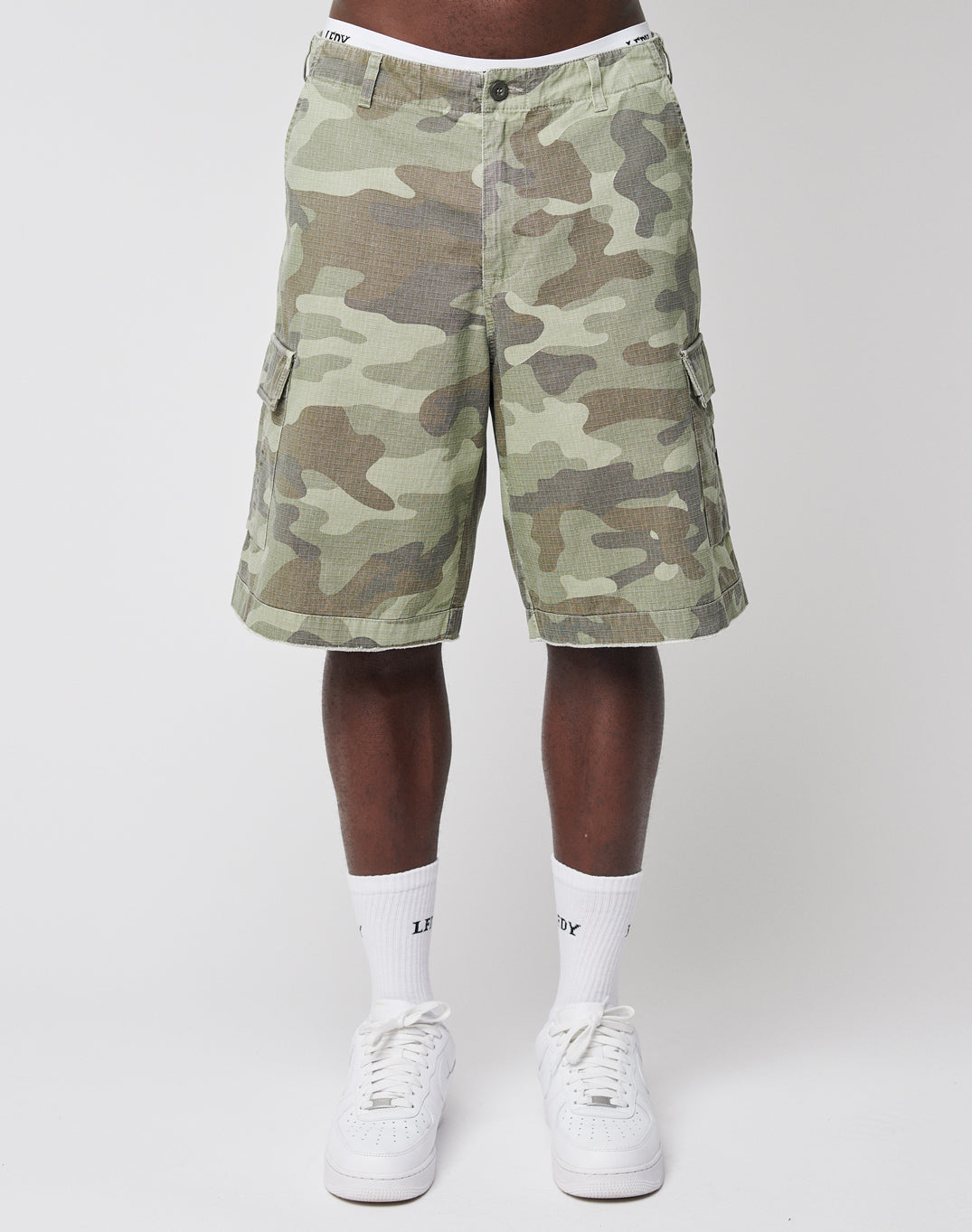 A person wearing LIVE FAST DIE YOUNG Army Shorts in green camouflage with klassischen Cargotaschen, white socks with LEGACY text, and white sneakers stands against a plain white background. Upper body not visible.
