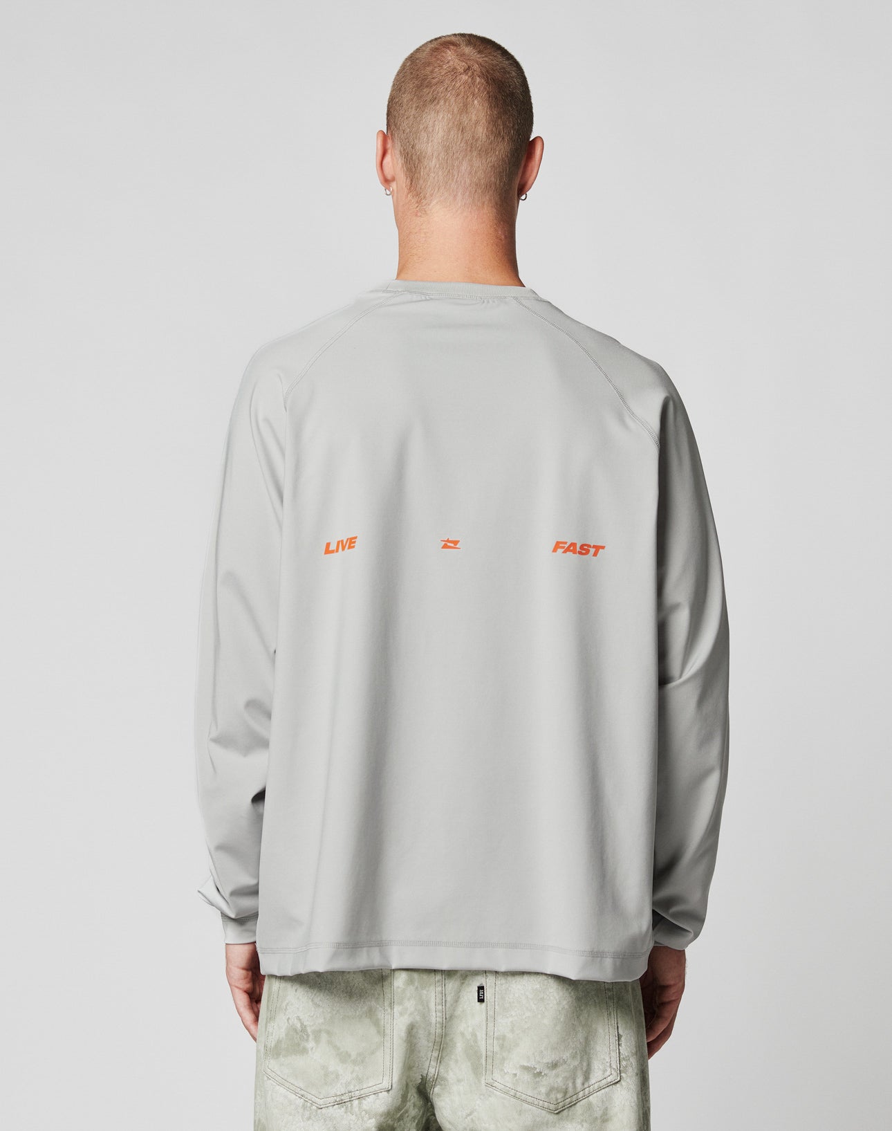 A person facing away wears the LFDY Sports Longsleeve by LIVE FAST DIE YOUNG in light gray with Raglan sleeves, oversize fit, and LIVE 2 FAST in small orange text on the back, paired with light green pants against a plain background.