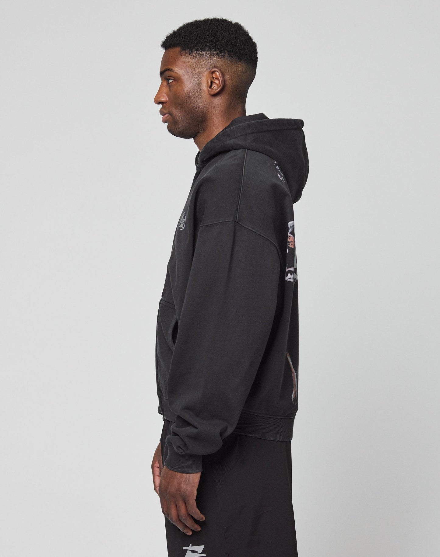 A man stands in profile against a plain background, wearing the LIVE FAST DIE YOUNG LFDY Canine Hooded Zip—an oversize black hooded sweatshirt with silver print and black pants, made from a cotton-polyester blend. His hands rest relaxed as he looks ahead.