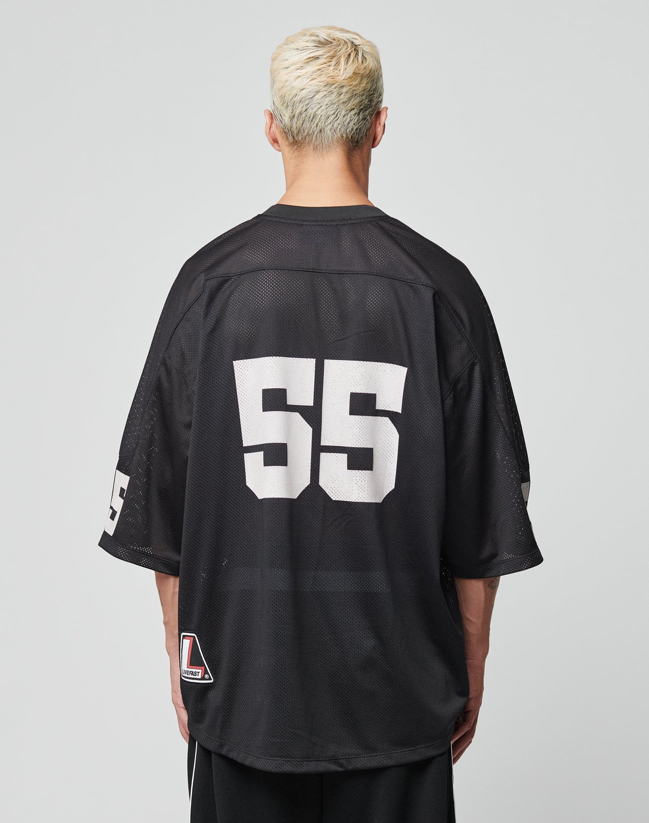 A person with short, bleached blonde hair is seen from the back wearing an oversized black LIVE FAST DIE YOUNG LFDY Mesh Jersey with large white 55 on the back and smaller 55 on the sleeve, set against a plain light gray background.