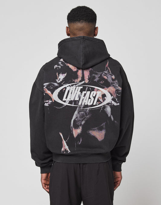 A person faces away from the camera in an oversize black LFDY Canine Hooded Zip by LIVE FAST DIE YOUNG, made from Baumwolle Polyester. It features snarling dog graphics and bold silver LIVE FAST text on the back, hood up. | model