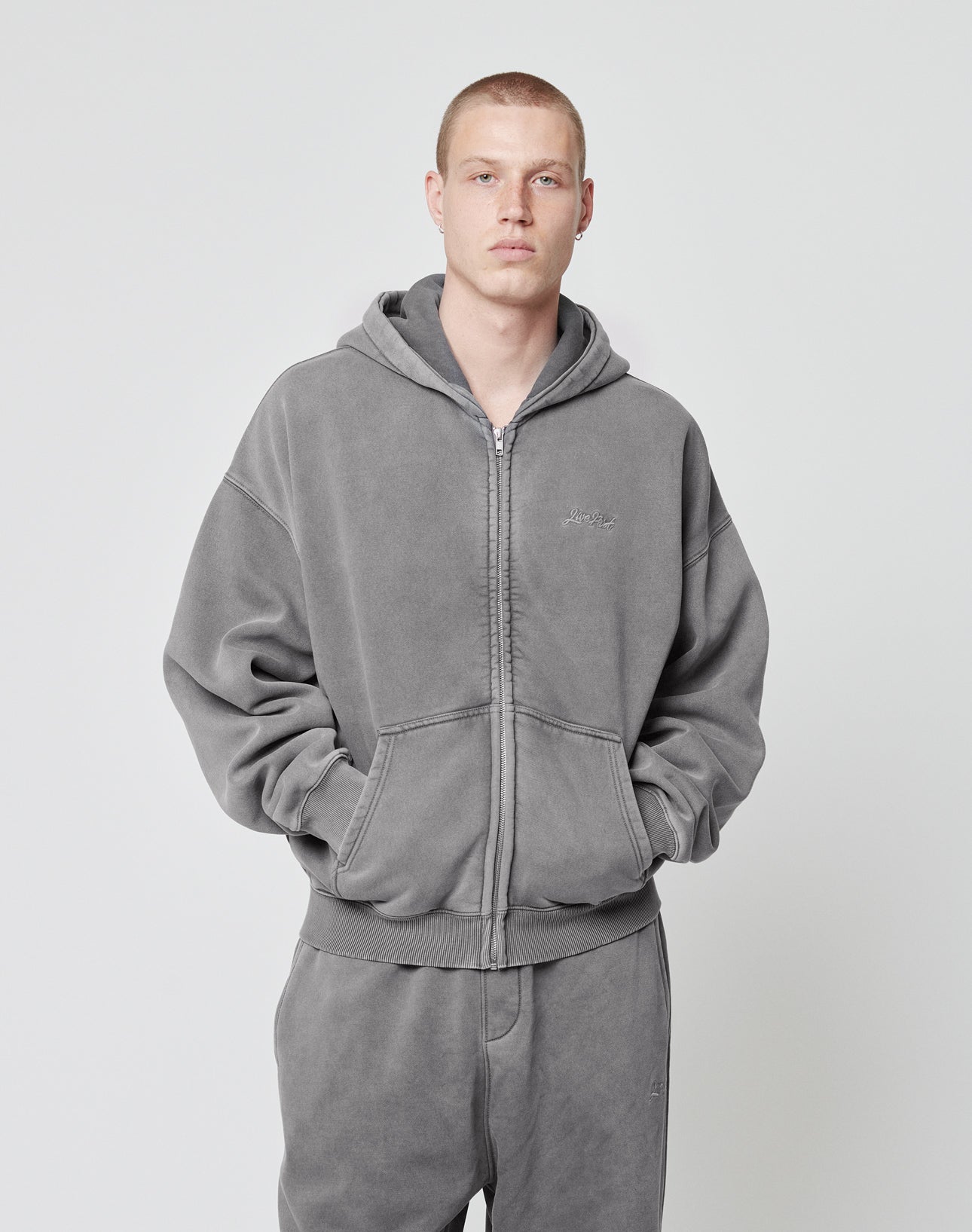 A person with short hair wears a matching grey LIVE FAST DIE YOUNG Basic Script Hooded Zip and sweatpants with Logo-Stick, facing the camera with hands in hoodie pockets against a plain light background.