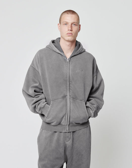A person with short hair wears a matching grey LIVE FAST DIE YOUNG Basic Script Hooded Zip and sweatpants with Logo-Stick, facing the camera with hands in hoodie pockets against a plain light background.