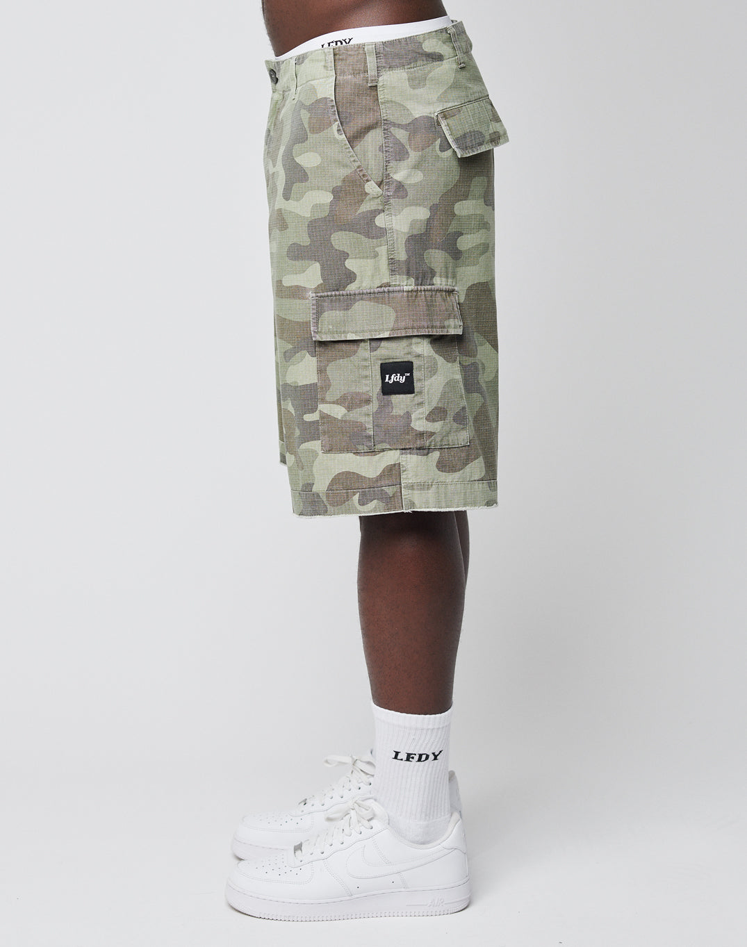 A person wearing Army Shorts from LIVE FAST DIE YOUNG, white LFDY socks, and white Nike sneakers stands sideways against a plain white background for a classic vintage look.