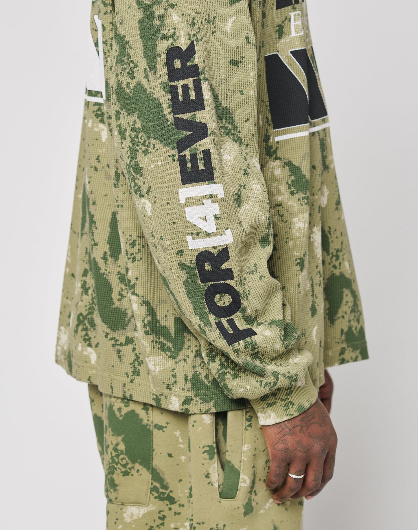A person wears the LFDY Racing Waffle Longsleeve by LIVE FAST DIE YOUNG—an oversize, green camouflage Baumwolle top with FOREVER in black and white on the sleeve. Their hand rests at their side against a plain background.