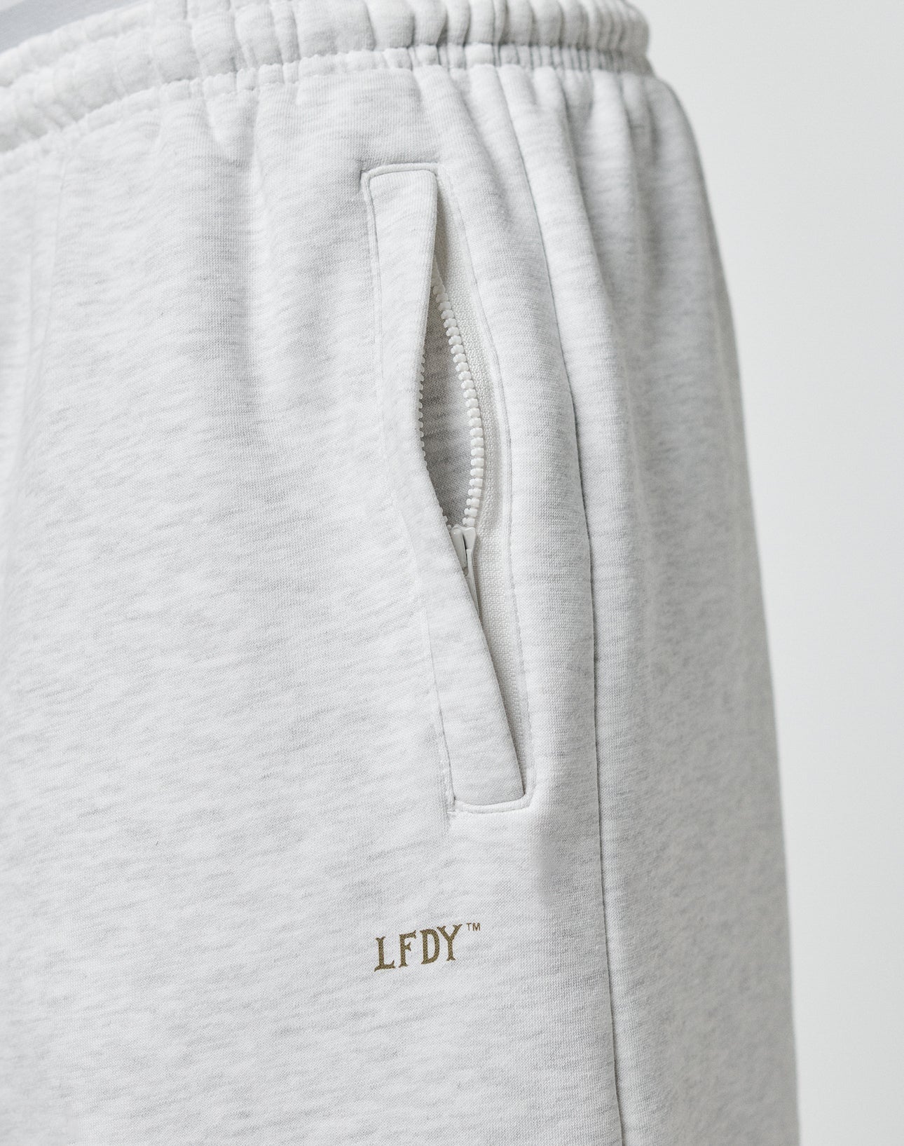 Close-up of the Basic Loose Jogger by LIVE FAST DIE YOUNG in light gray, showing a pocket with a white zipper and elastic waistband, plus gold embroidered LFDY logo near the pocket.