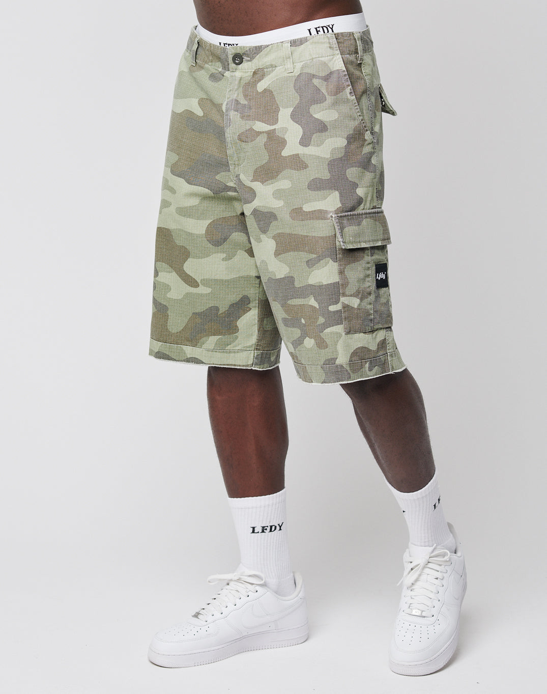 A person wears LIVE FAST DIE YOUNG Army Shorts with green camouflage, side pockets, and a black logo patch, paired with white LEDY socks and white sneakers. Only the lower body is visible, set against a plain background.