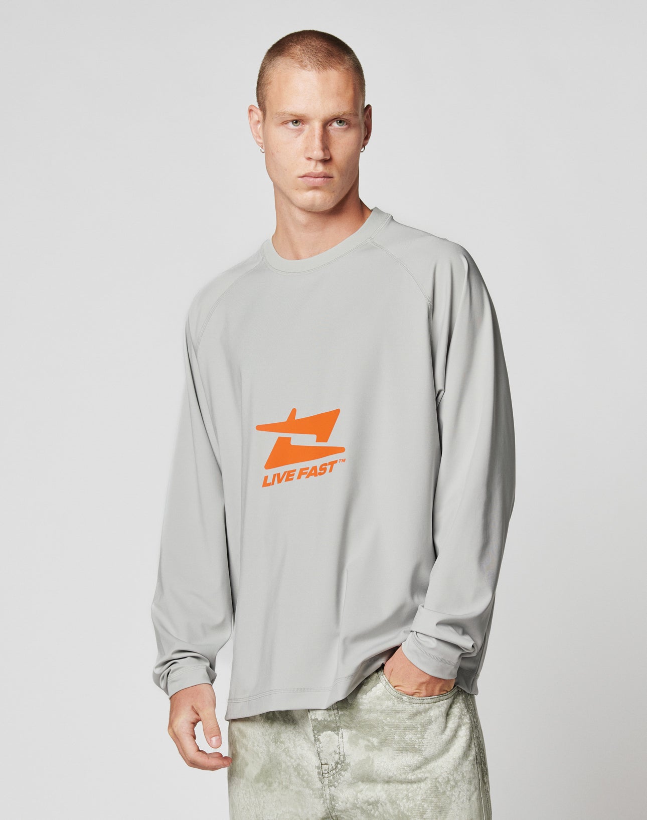 A man with short hair wears the light gray LFDY Sports Longsleeve by LIVE FAST DIE YOUNG, featuring Raglan sleeves and an orange LIVE FAST graphic, paired with light pants. He stands against a plain, light background, looking to the side.