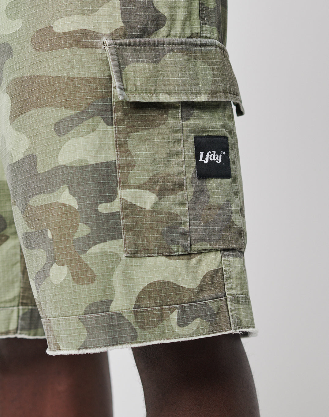 A close-up shows a person wearing Army Shorts by LIVE FAST DIE YOUNG, styled in green with a vintage look, featuring cargo pockets and a side pocket adorned with a black Lfdy™ patch label against a plain background.