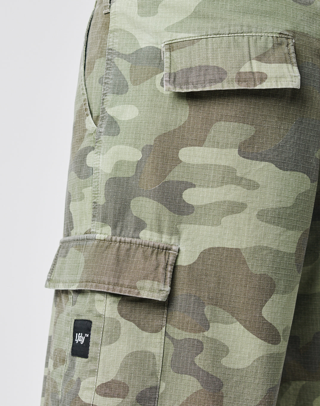 Close-up of LIVE FAST DIE YOUNG Army Shorts in a green, brown, and beige camo pattern. Features multiple cargo pockets, including a flap with a small black and white label, enhancing their vintage style.
