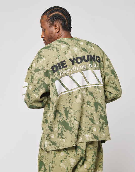 A man with cornrows faces away from the camera, wearing an oversized green camo LFDY Racing Waffle Longsleeve by LIVE FAST DIE YOUNG. The back features DIE YOUNG Everything in it and a bold white graphic. Made from 100% Baumwolle.