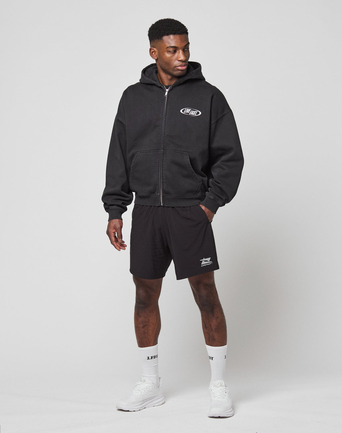 A man stands against a plain background wearing the LFDY Canine Hooded Zip by LIVE FAST DIE YOUNG, black shorts, white crew socks, and white sneakers. He has one hand in his pocket and looks to the side.