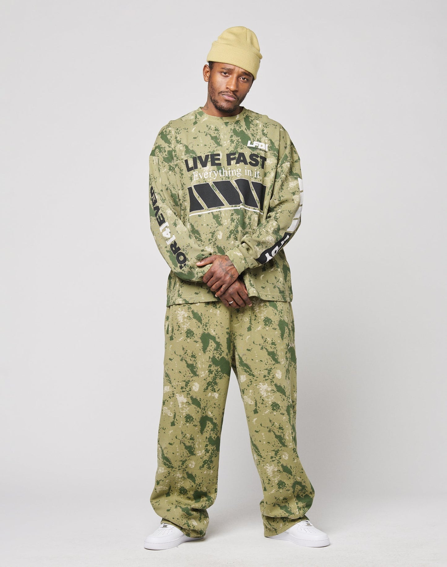 A man in a yellow beanie, LIVE FAST DIE YOUNG Camo Wide Jogger set, and white sneakers stands against a plain light background. His sweatshirt features the LIVE FAST slogan.