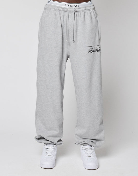 A person in light gray Wide Cuff Jogger sweatpants by LIVE FAST DIE YOUNG, featuring LIVE FAST on the waistband and thigh, plus white sneakers, stands against a plain white backdrop.