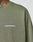 Close-up of a person wearing the LIVE FAST DIE YOUNG Basic Longsleeve in olive green, featuring a white logo print on the chest. Oversize fit. 100% Baumwolle. Part of the neck and a tattoo are visible.
