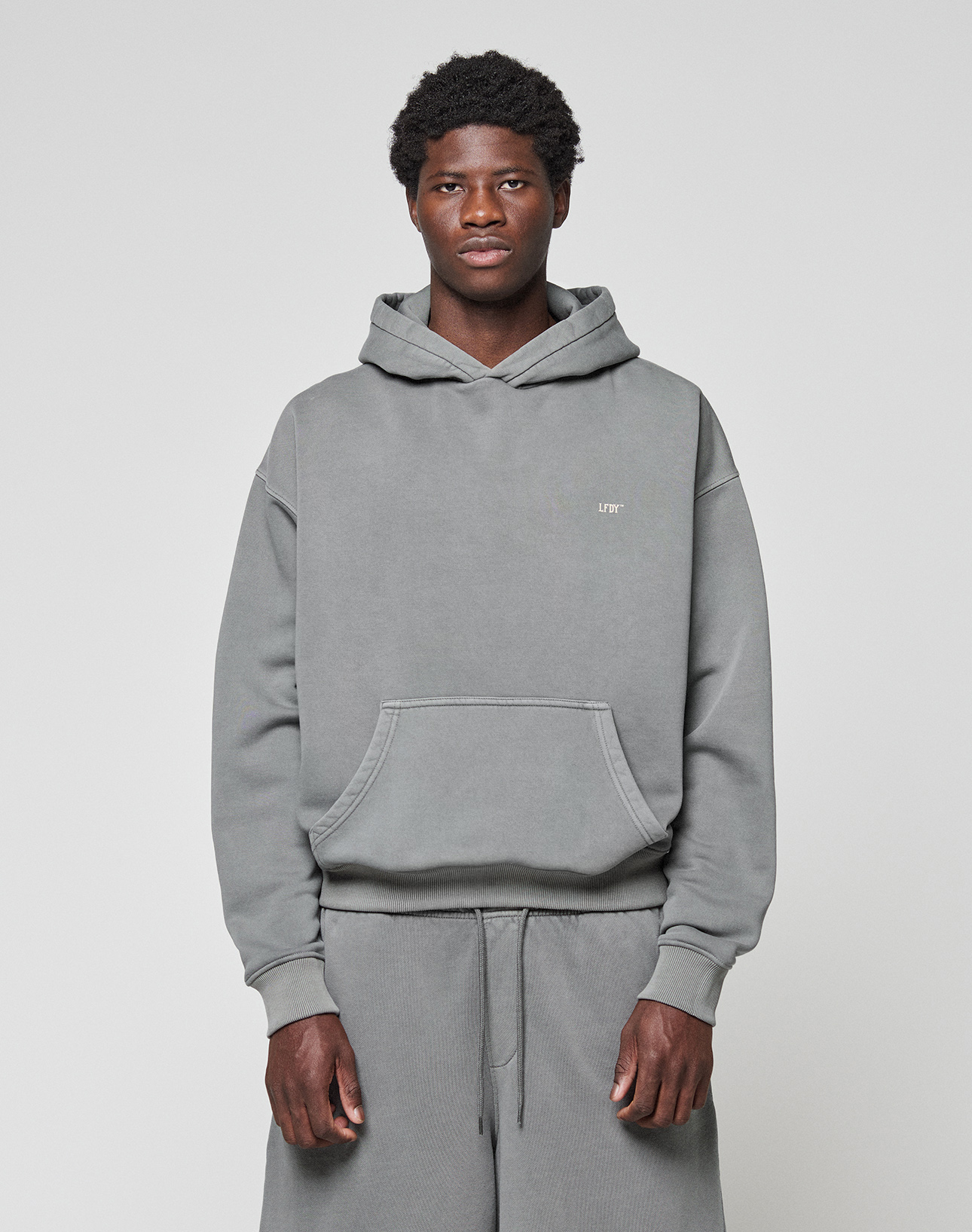 A man stands against a plain light gray background, wearing the LIVE FAST DIE YOUNG Basic Evidence Hooded gray hoodie with front pocket and matching sweatpants, looking directly at the camera with a neutral expression.
