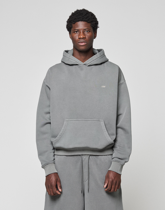 A man stands against a plain light gray background, wearing the LIVE FAST DIE YOUNG Basic Evidence Hooded gray hoodie with front pocket and matching sweatpants, looking directly at the camera with a neutral expression.
