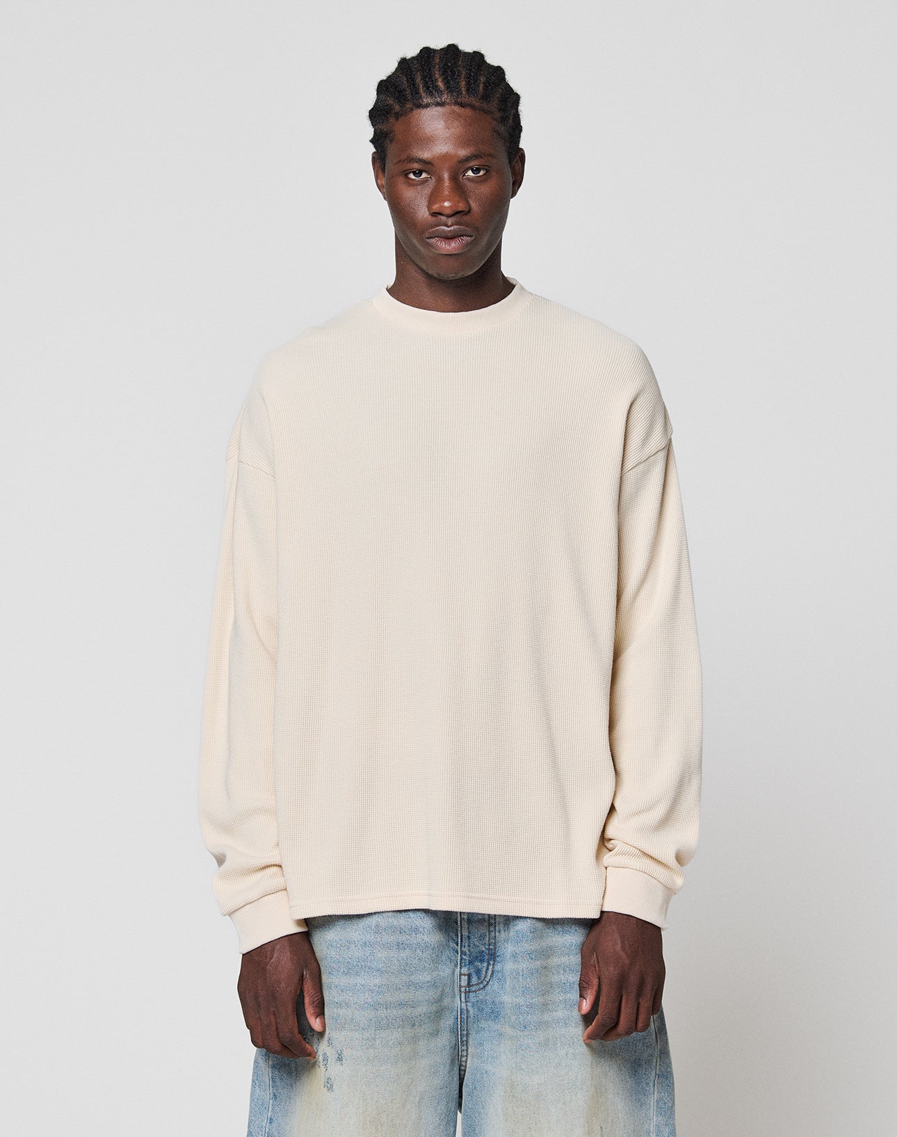 A man with braided hair wears the LIVE FAST DIE YOUNG Blank Waffle Longsleeve in cream, an oversized 100% cotton shirt, paired with light blue jeans, standing against a plain light gray background and looking at the camera. | model