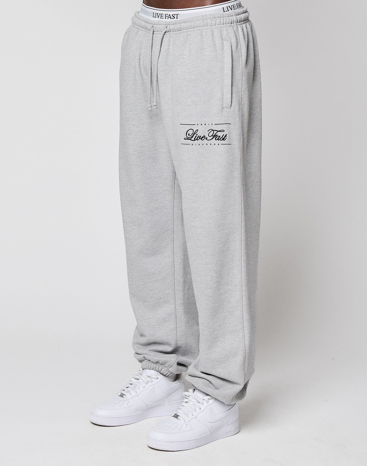 A model wears LIVE FAST DIE YOUNG Wide Cuff Jogger in light gray with "Live Fast" text on the left thigh and a comfy elastic waistband, styled with white sneakers. The photo shows the joggers waist to ankles against a plain white background.
