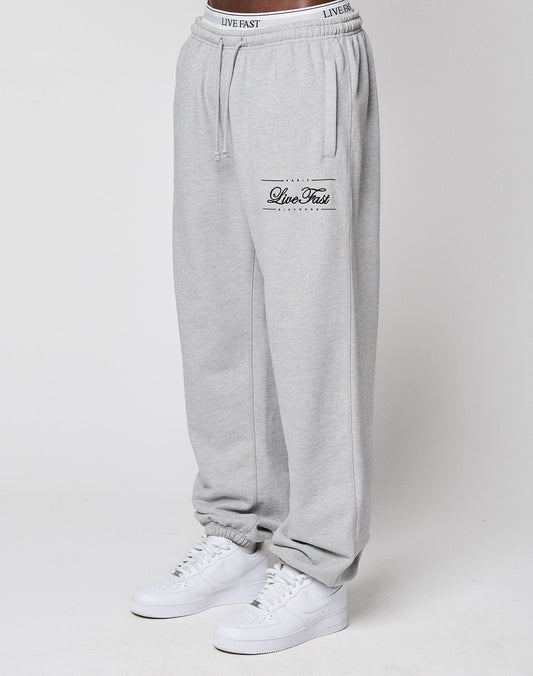 A model wears LIVE FAST DIE YOUNG Wide Cuff Jogger in light gray with "Live Fast" text on the left thigh and a comfy elastic waistband, styled with white sneakers. The photo shows the joggers waist to ankles against a plain white background.
