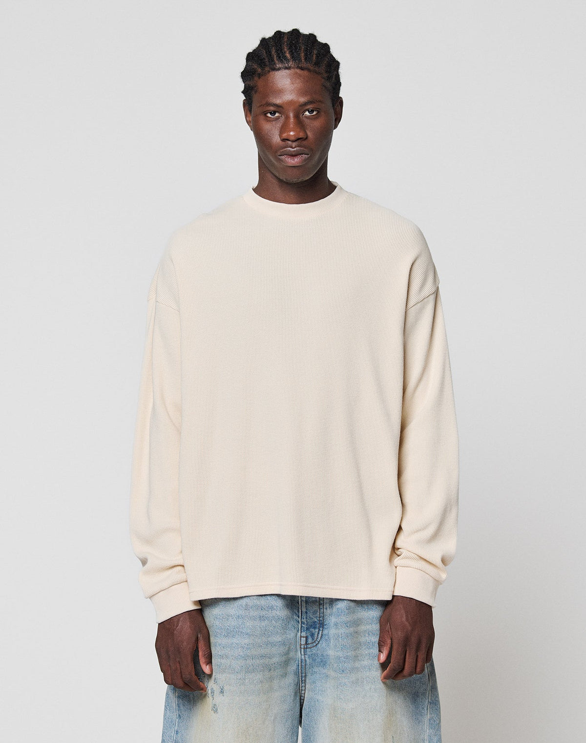 A man with braided hair wears the LIVE FAST DIE YOUNG Blank Waffle Longsleeve in cream, an oversized 100% cotton shirt, paired with light blue jeans, standing against a plain light gray background and looking at the camera. | model