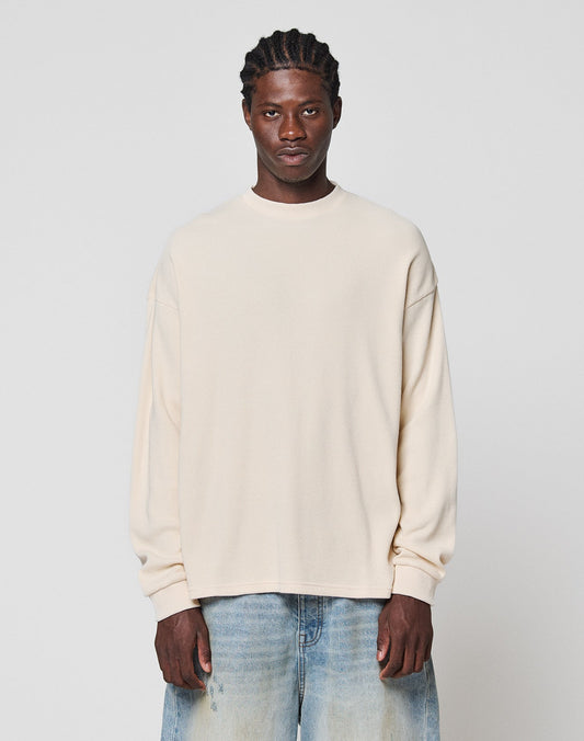 A man with braided hair wears the LIVE FAST DIE YOUNG Blank Waffle Longsleeve in cream, an oversized 100% cotton shirt, paired with light blue jeans, standing against a plain light gray background and looking at the camera. | model