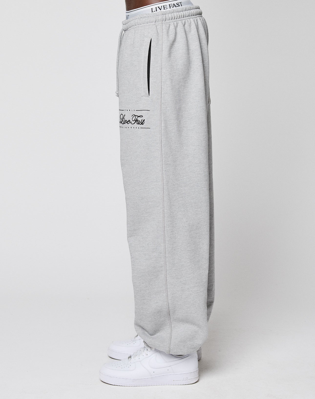 A person stands sideways in light gray Wide Cuff Jogger sweatpants by LIVE FAST DIE YOUNG, featuring a branded elastic waistband and pocket text, paired with white sneakers against a plain white background.