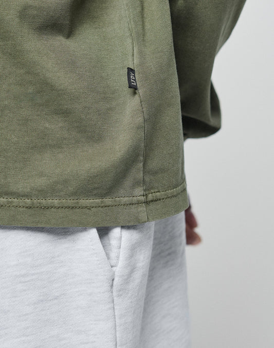 Close-up of a person wearing the LIVE FAST DIE YOUNG Basic Longsleeve in light gray (100% Baumwolle) with a pocket, olive green oversize fit, and a small black side-seam tag. Background is plain and light-colored.